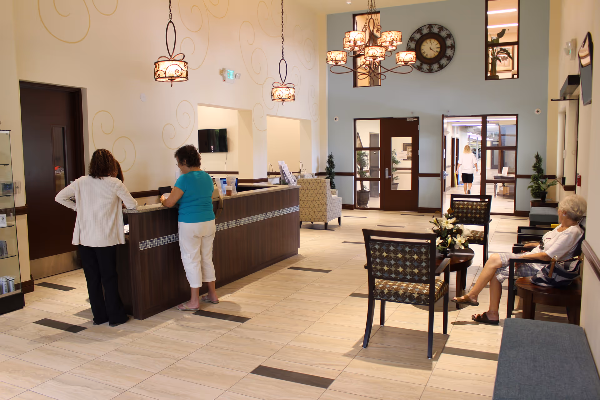 Bright senior living lobby with a reception desk where two people stand and a seating area with a woman seated.