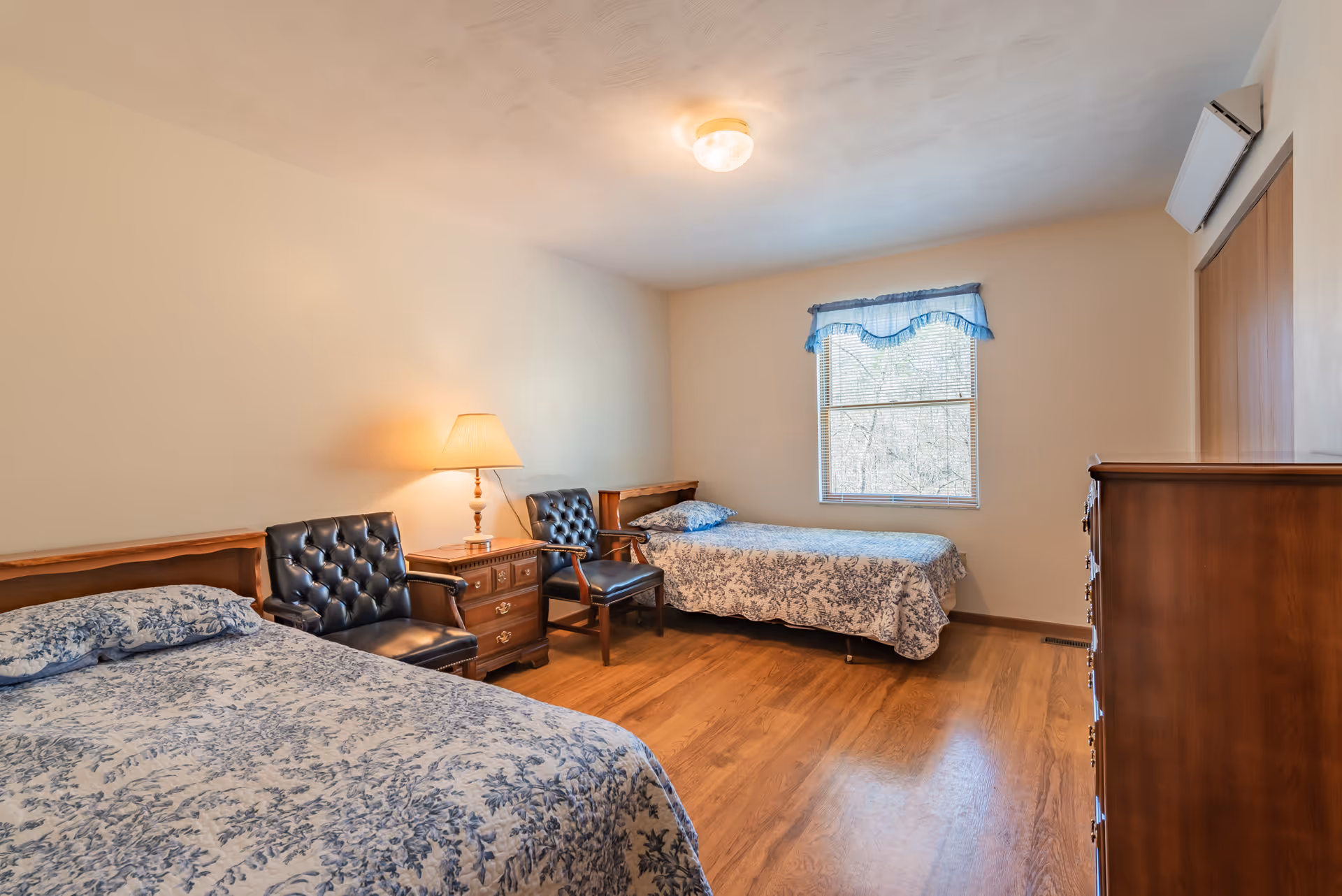 A simple bedroom with two twin beds covered in blue and white floral bedspreads. Between the beds is a wooden nightstand with a lamp on it. Two black leather chairs are positioned next to the nightstand. There is a window with a blue valance letting in natural light, and a wooden dresser is visible on the right side of the room. The floor is wooden, and the walls are painted a light beige color.