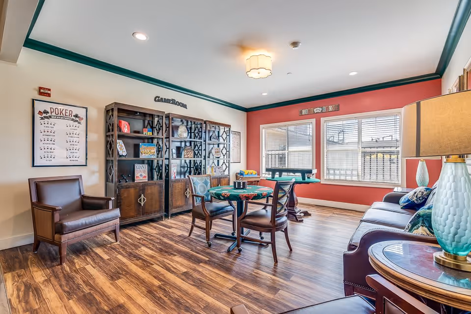 A bright game room in a senior living facility with wooden flooring, a round table with chairs, a sofa with colorful pillows, a side table with a lamp, and large windows letting in natural light. There are bookshelves with games and decorations, and a poster about poker on the wall.