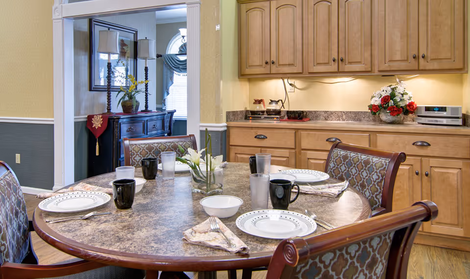 Round dining table set with plates, cups, and a floral centerpiece in a dining area adjoining kitchen cabinets.