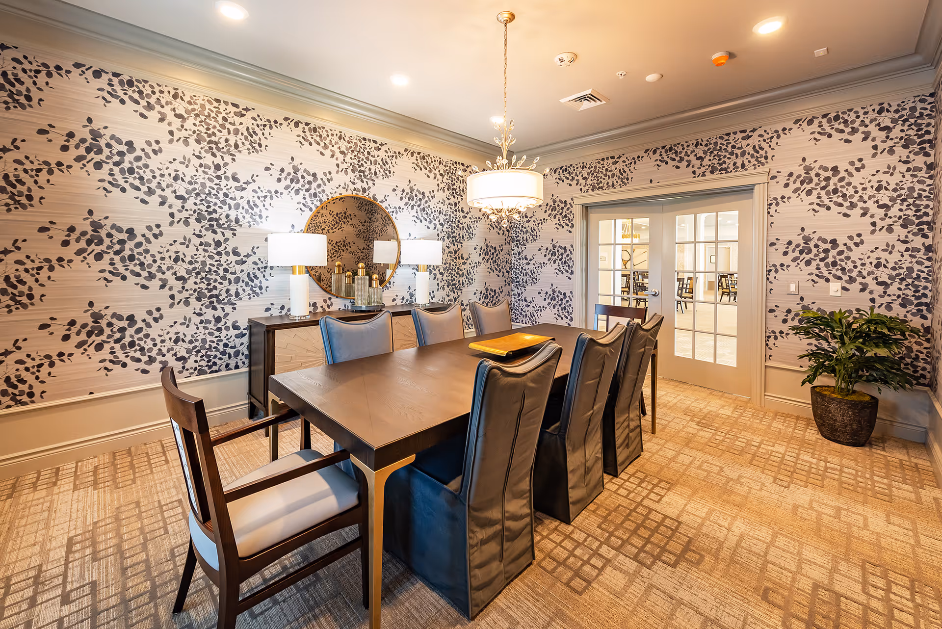 Elegant dining room with a long wooden table surrounded by upholstered chairs, patterned wallpaper, a round mirror, lamps, and a chandelier.