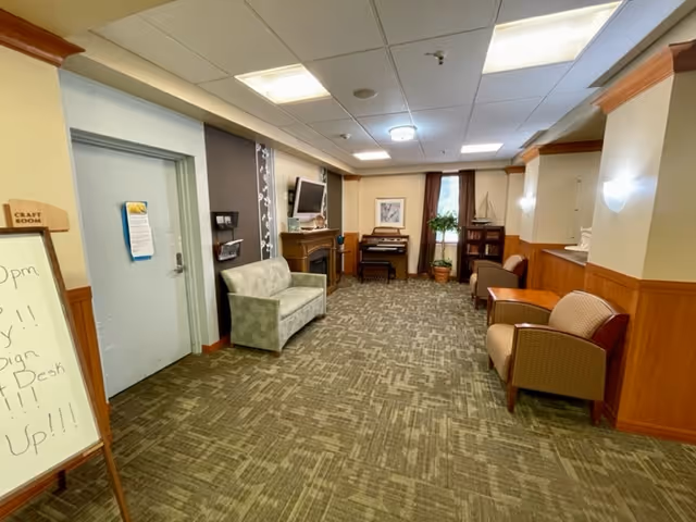 Interior view of a senior living facility common area with carpeted floor, beige walls with wood paneling, a small couch, armchairs, a piano, a TV mounted above a fireplace, and a whiteboard sign near a door labeled 'Craft Room'.
