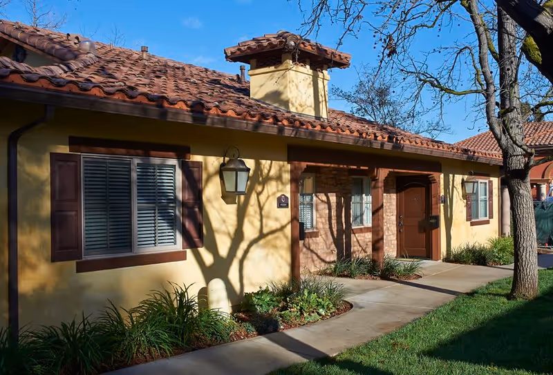 Exterior view of a single-story building with a yellow stucco facade, brown wooden shutters, and a red tile roof. There is a pathway leading to a brown door, with plants and grass along the side. A tree casts shadows on the building and the sky is clear and blue.