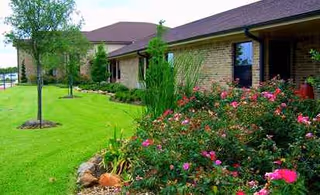 Exterior view of a single-story brick senior facility with manicured lawn and flowering shrubs.
