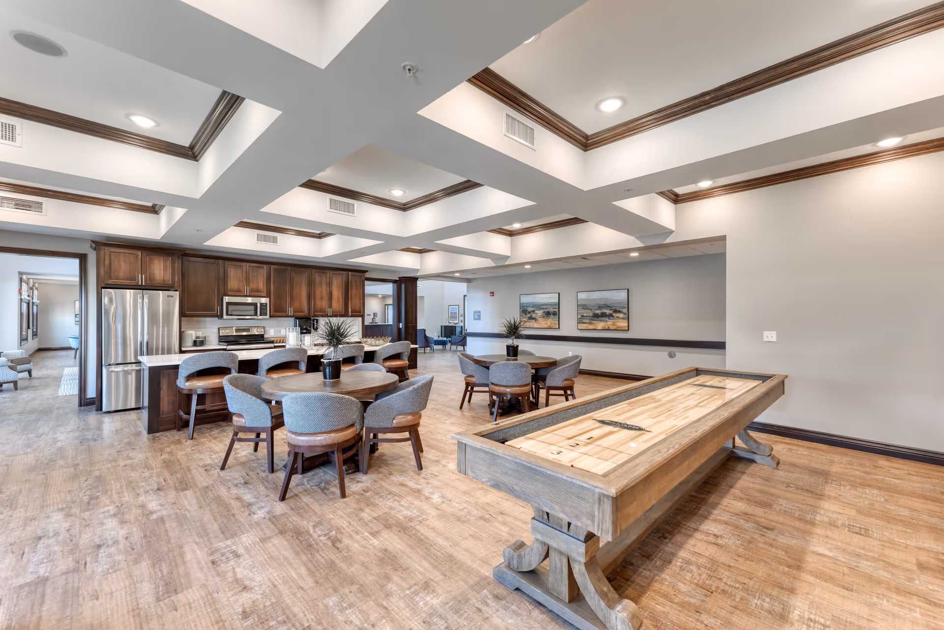 A spacious senior living common area featuring a modern kitchen with dark wood cabinets and stainless steel appliances, round tables with upholstered chairs, and a wooden shuffleboard table. The room has a coffered ceiling with recessed lighting and light wood flooring.