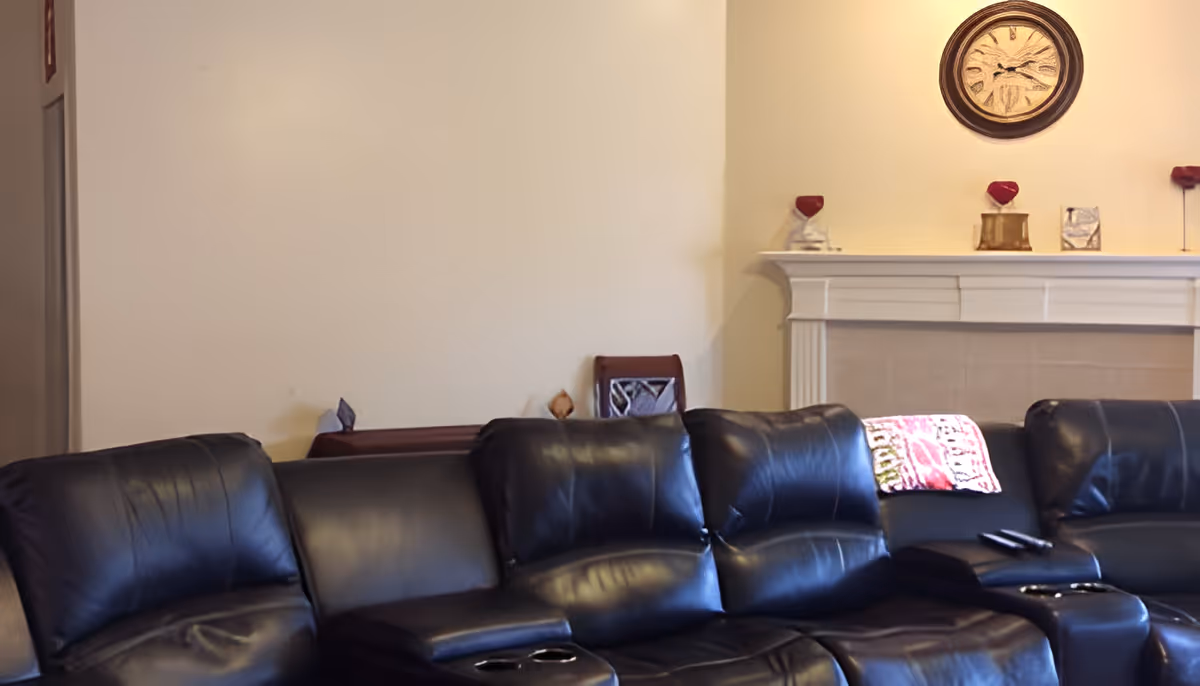 A living room with a black leather sectional sofa facing away from a white fireplace mantel. The mantel is decorated with small decorative items and a round wall clock hangs above it. A colorful throw blanket is draped over one section of the sofa, and two remote controls rest on the armrest.