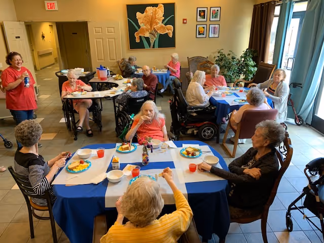 Seniors seated at several tables in a communal dining room eating and socializing.