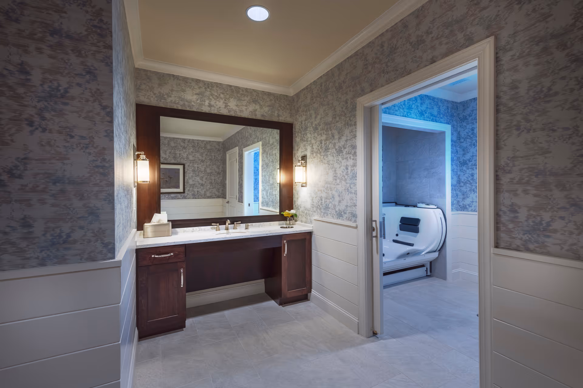 Well-lit accessible bathroom with a large vanity and mirror, and a walk-in bathing unit visible through an open doorway.