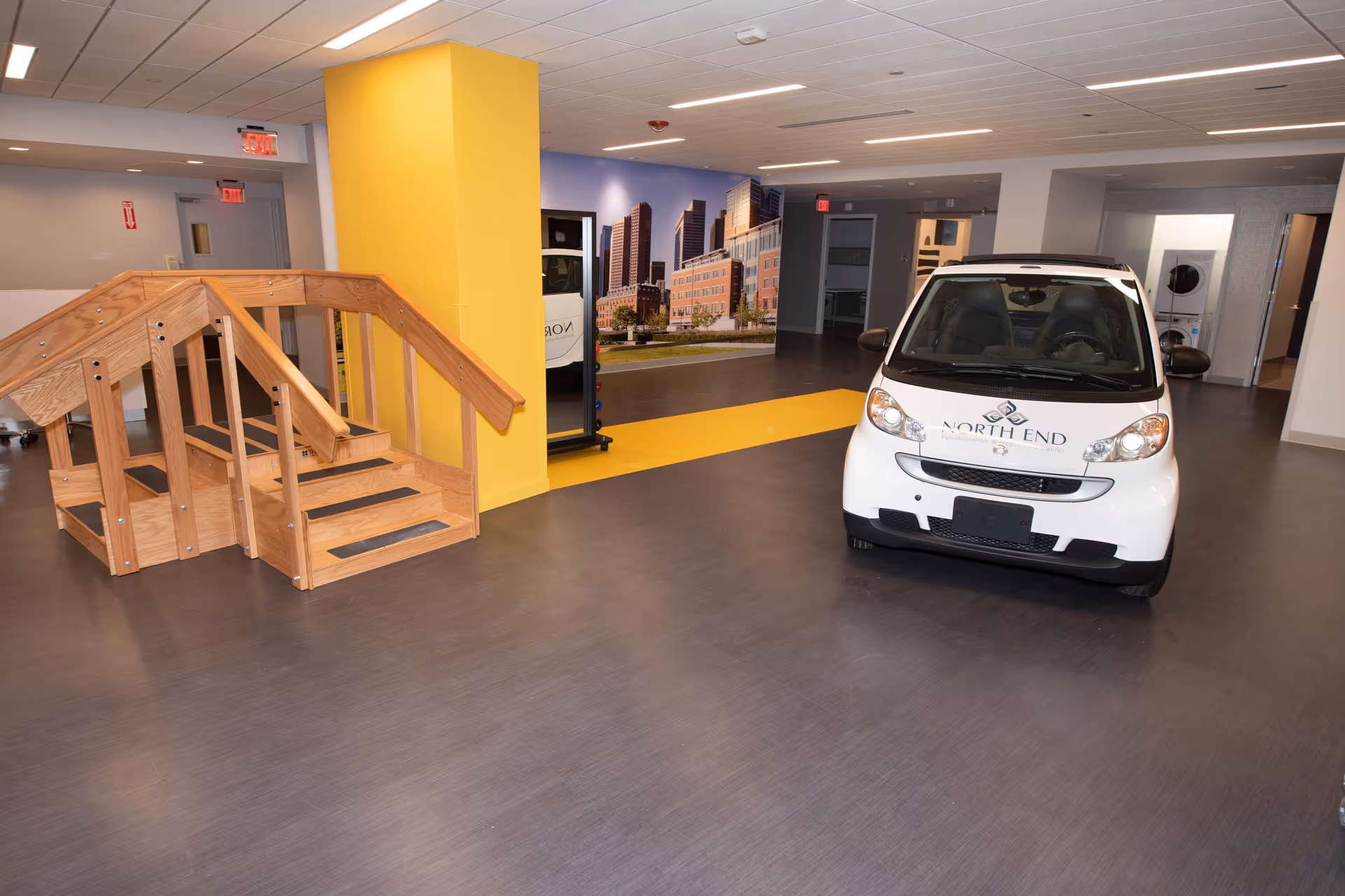 Interior of a rehabilitation facility showing a small white North End branded car, wooden therapy stairs, a yellow column, and a city mural on the wall.