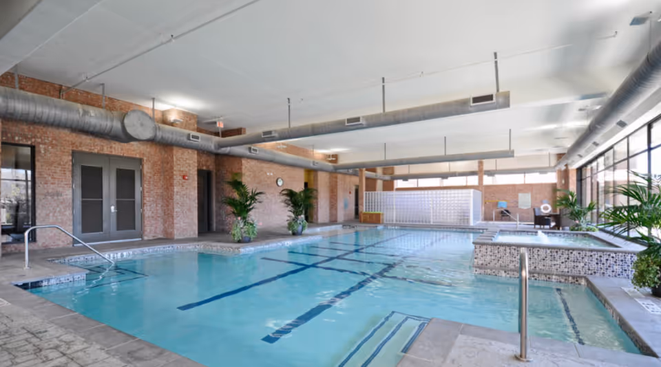 Indoor lap pool with tiled hot tub, plants, and large windows in a bright brick-walled recreation room.