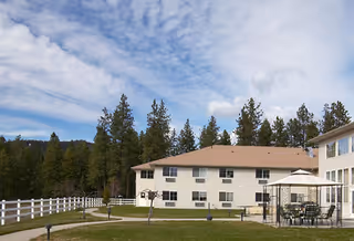 Outdoor view of a senior living facility with a two-story beige building, a white fence, patio area with tables and chairs under an umbrella, surrounded by green grass and tall pine trees under a partly cloudy sky.