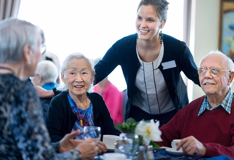 A caregiver smiles as elderly residents sit around a dining table with cups and a flower centerpiece.