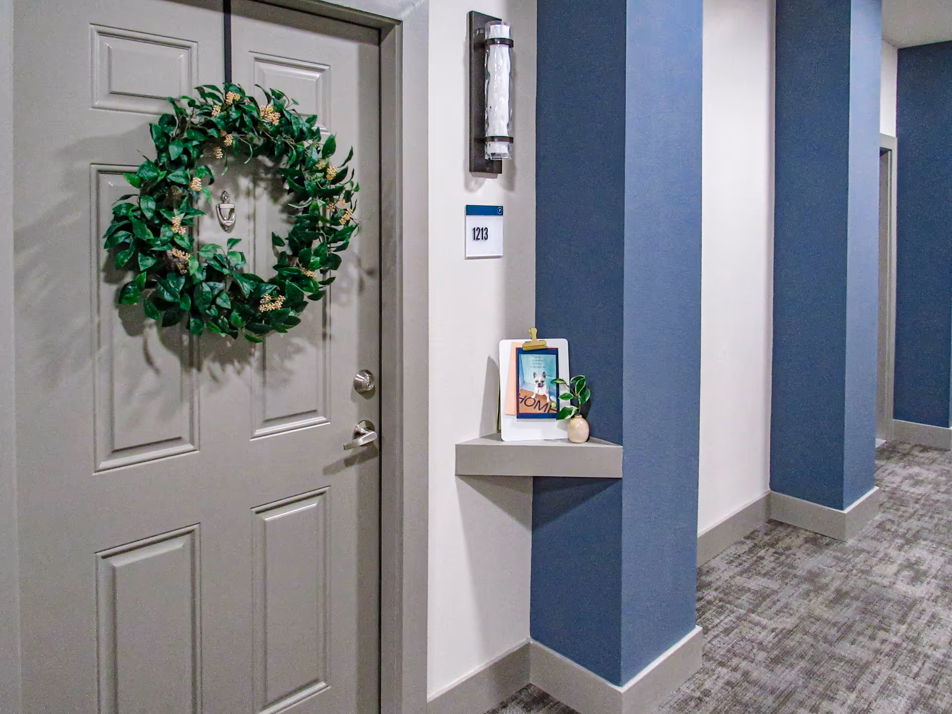 Interior hallway of a senior living facility with a closed door decorated with a green leafy wreath. The door has a silver handle and a door knocker. Next to the door is a small corner shelf holding a framed picture and a small plant. The walls are painted white and blue, and the floor is carpeted in a gray pattern. A room number plaque labeled 1213 is mounted on the wall above the shelf.
