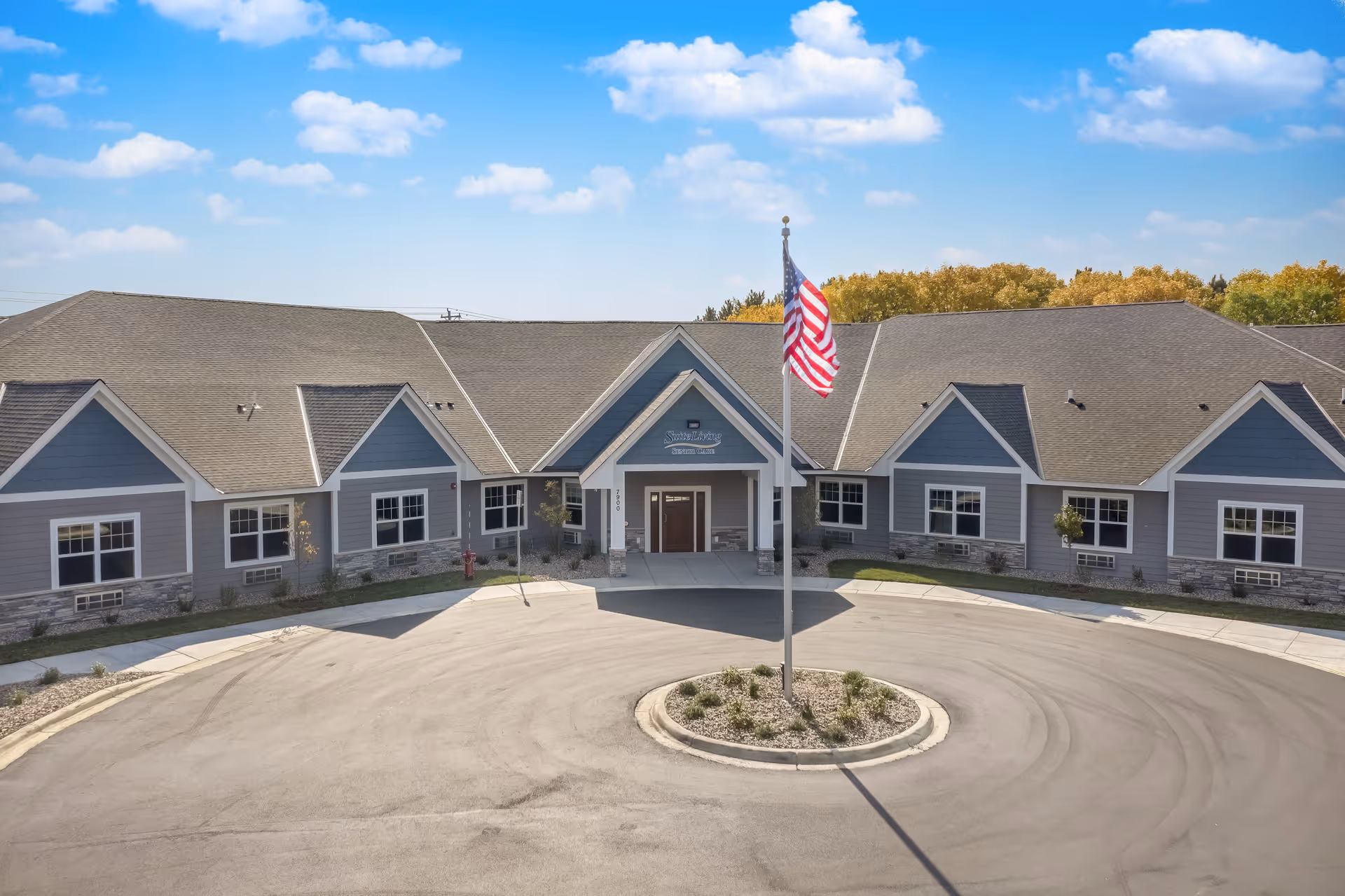 Front exterior view of Suite Living of Inver Grove Heights, a single-story building with gray siding and a peaked roof. An American flag is prominently displayed on a flagpole in the circular driveway in front of the entrance. The sky is blue with scattered clouds.