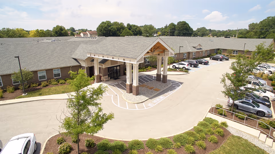 Exterior view of Harbour Manor & The Lodge showing the main entrance with a covered drop-off area, surrounded by parking spaces and landscaped greenery under a partly cloudy sky.