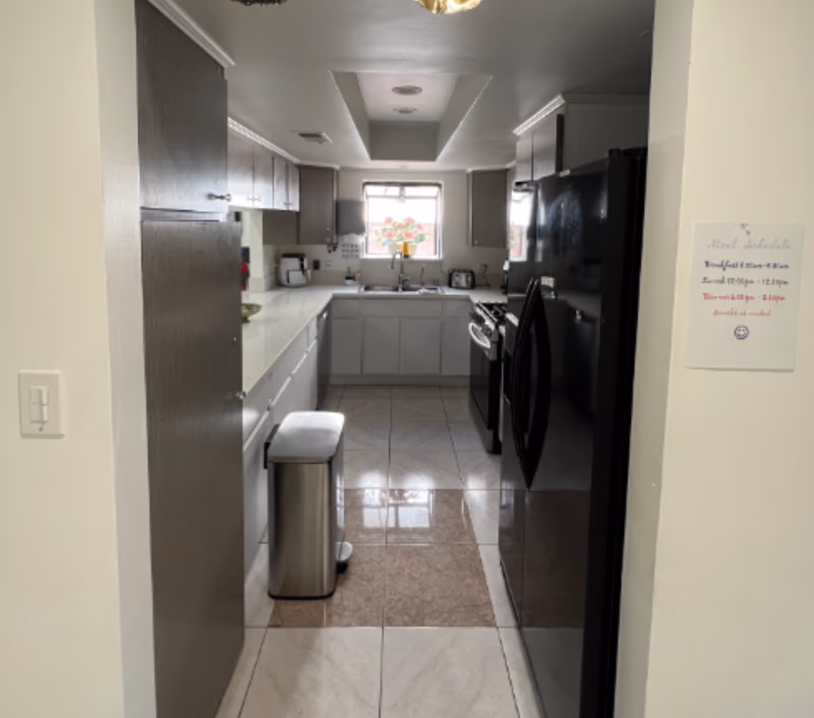 A clean, modern kitchen with white countertops and cabinets, dark upper cabinets, a black refrigerator, a stove, a stainless steel trash can, and a window above the sink with a flower decoration. A meal schedule is posted on the wall to the right.