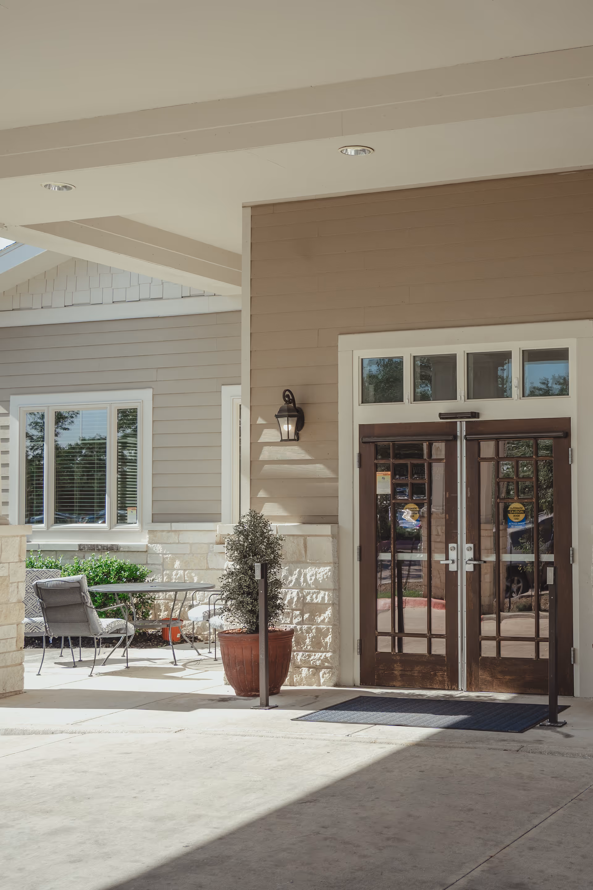 Entrance area of a senior living facility with double wooden doors featuring glass panels, a small outdoor seating area with metal chairs and a table, potted plants, and beige siding with stone accents on the building exterior.