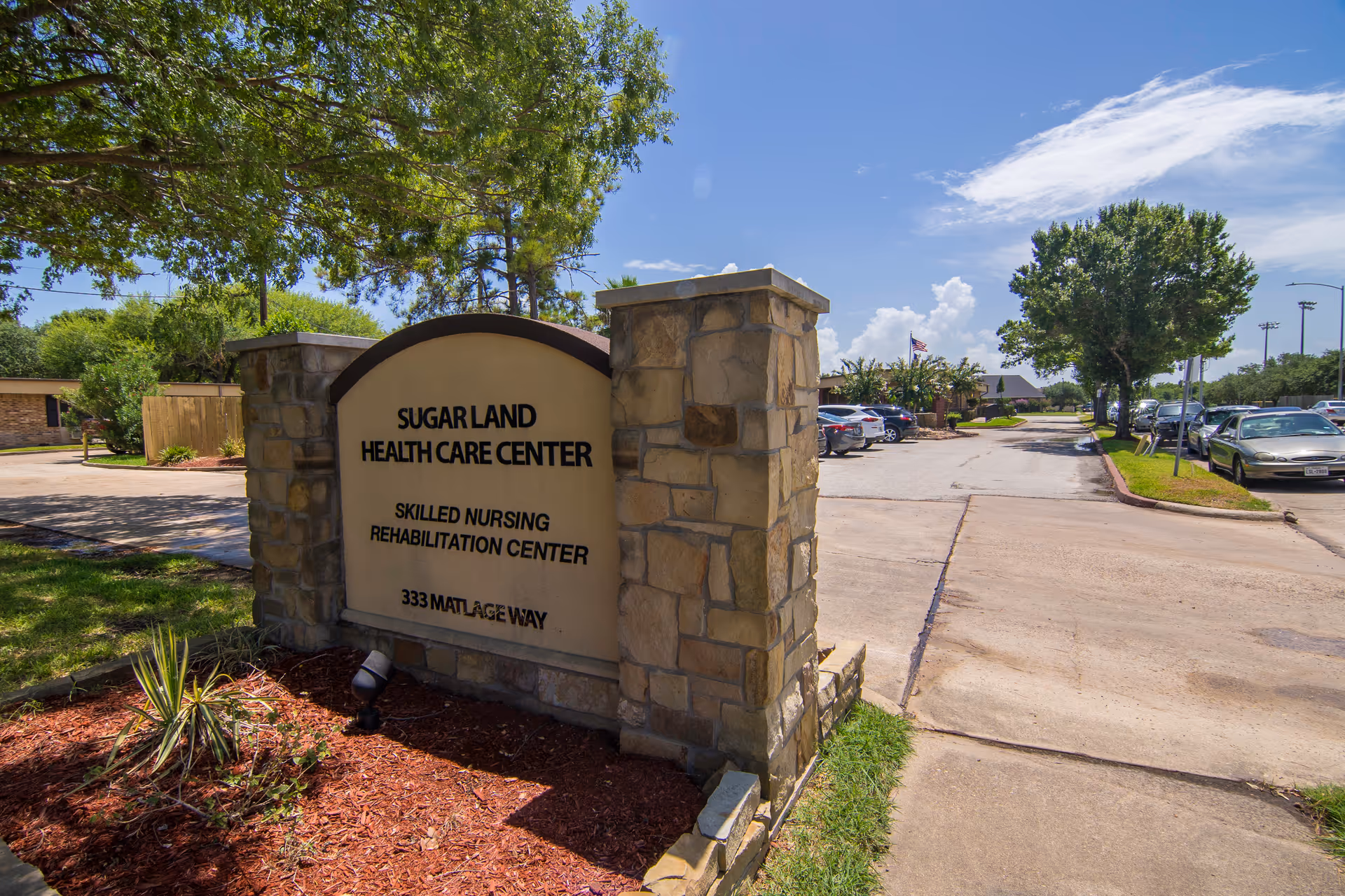 Stone sign for Sugar Land Health Care Center, a skilled nursing rehabilitation center, located at 333 Matlage Way, with a parking lot and trees in the background under a blue sky with some clouds.