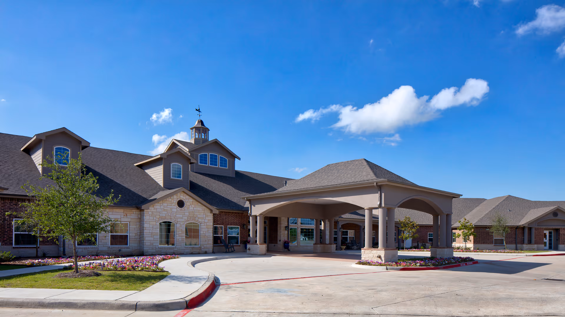 Exterior view of Twin Rivers Assisted Living and Memory Care facility showing a large covered entrance with columns, brick and stone building facade, a few small trees, and a clear blue sky with some clouds.