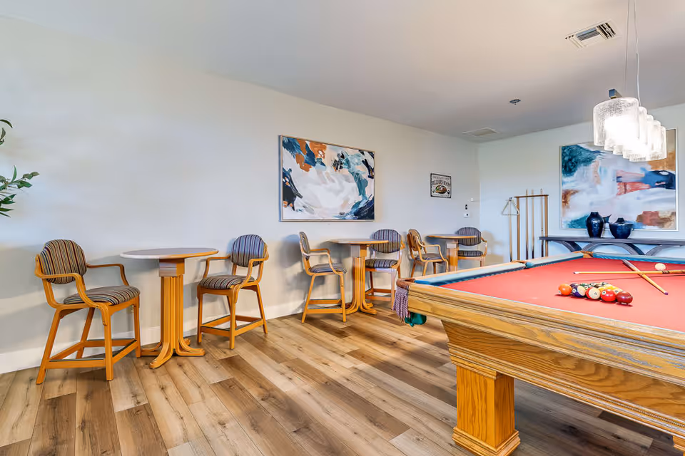 A recreational room with a red felt pool table in the foreground, several wooden tables with striped cushioned chairs along the wall, abstract paintings hanging on the walls, and a wooden floor.