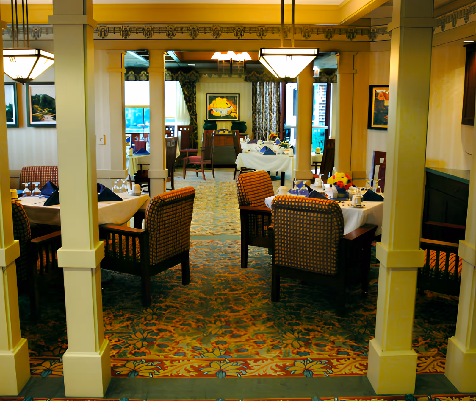 Interior view of a dining room in a senior living facility with multiple tables set with white tablecloths, napkins, glassware, and floral centerpieces. The room features patterned carpet, upholstered chairs, decorative columns, framed artwork on the walls, and large windows with curtains allowing natural light.