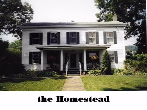 Front exterior view of a two-story white house with black shutters, a small porch with two chairs, surrounded by trees and greenery, with a lawn and a walkway leading to the entrance. The text 'the Homestead' is displayed below the image.