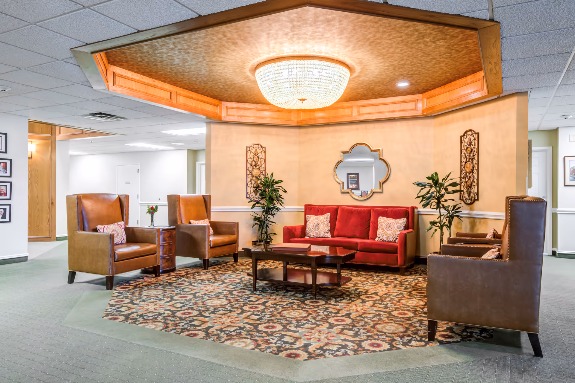 A cozy seating area in a senior living facility with a red sofa, four brown leather armchairs, a wooden coffee table, two potted plants, a decorative mirror on the wall, and a large chandelier overhead. The floor has a patterned area rug and the walls are painted beige with white trim.
