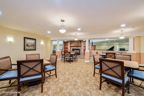 A spacious common area with multiple tables and chairs arranged on a patterned carpet. The room features warm beige walls, a stone fireplace in the background, and soft overhead lighting. There is an American flag near the fireplace and a framed picture on the left wall.