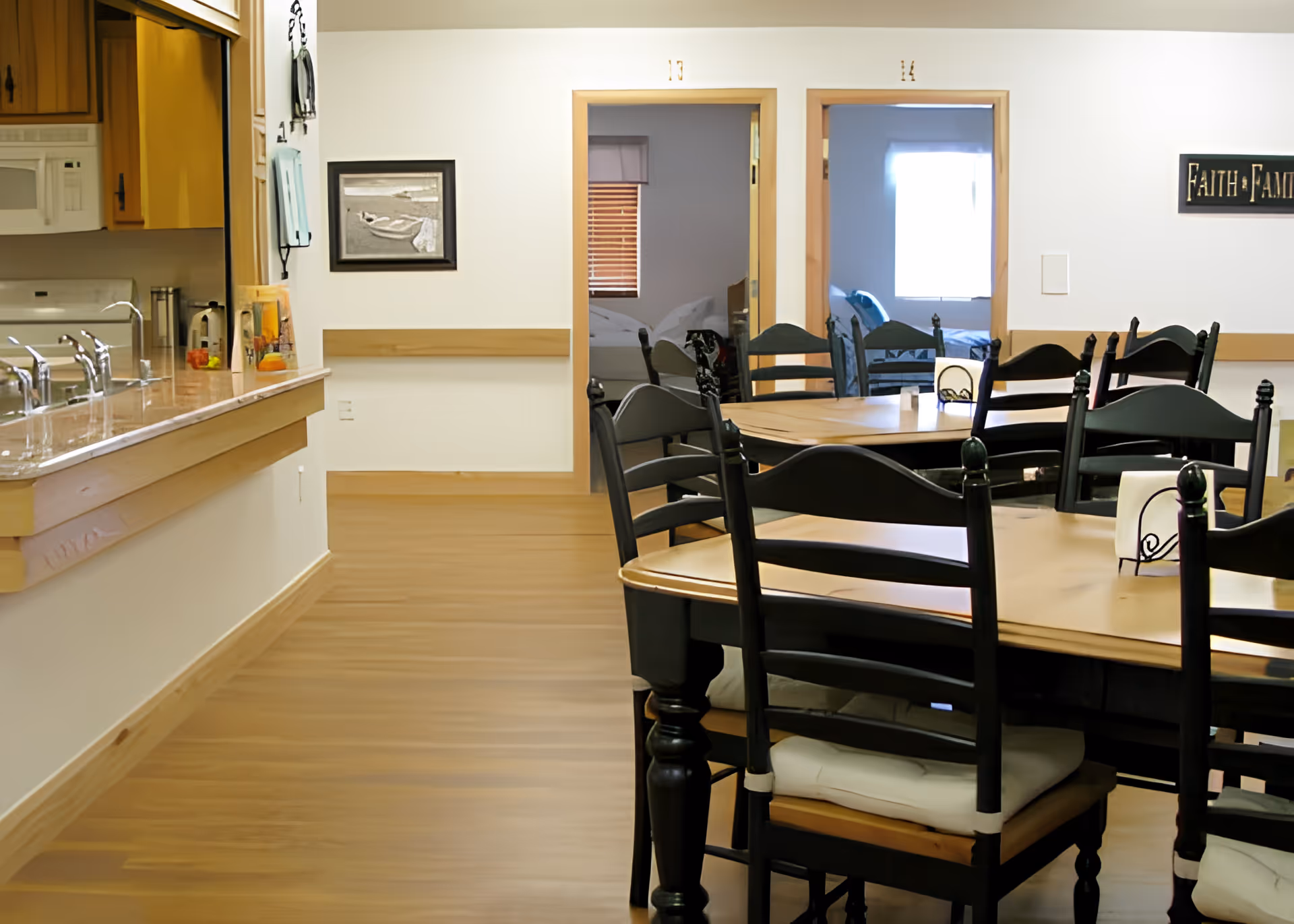 Interior view of a senior living facility dining area with wooden tables and black chairs with cushions. To the left is a kitchen counter with multiple sinks and a microwave. In the background, two doorways lead to bedrooms with beds visible inside. A framed picture and a sign that reads 'Faith Family' are mounted on the walls.