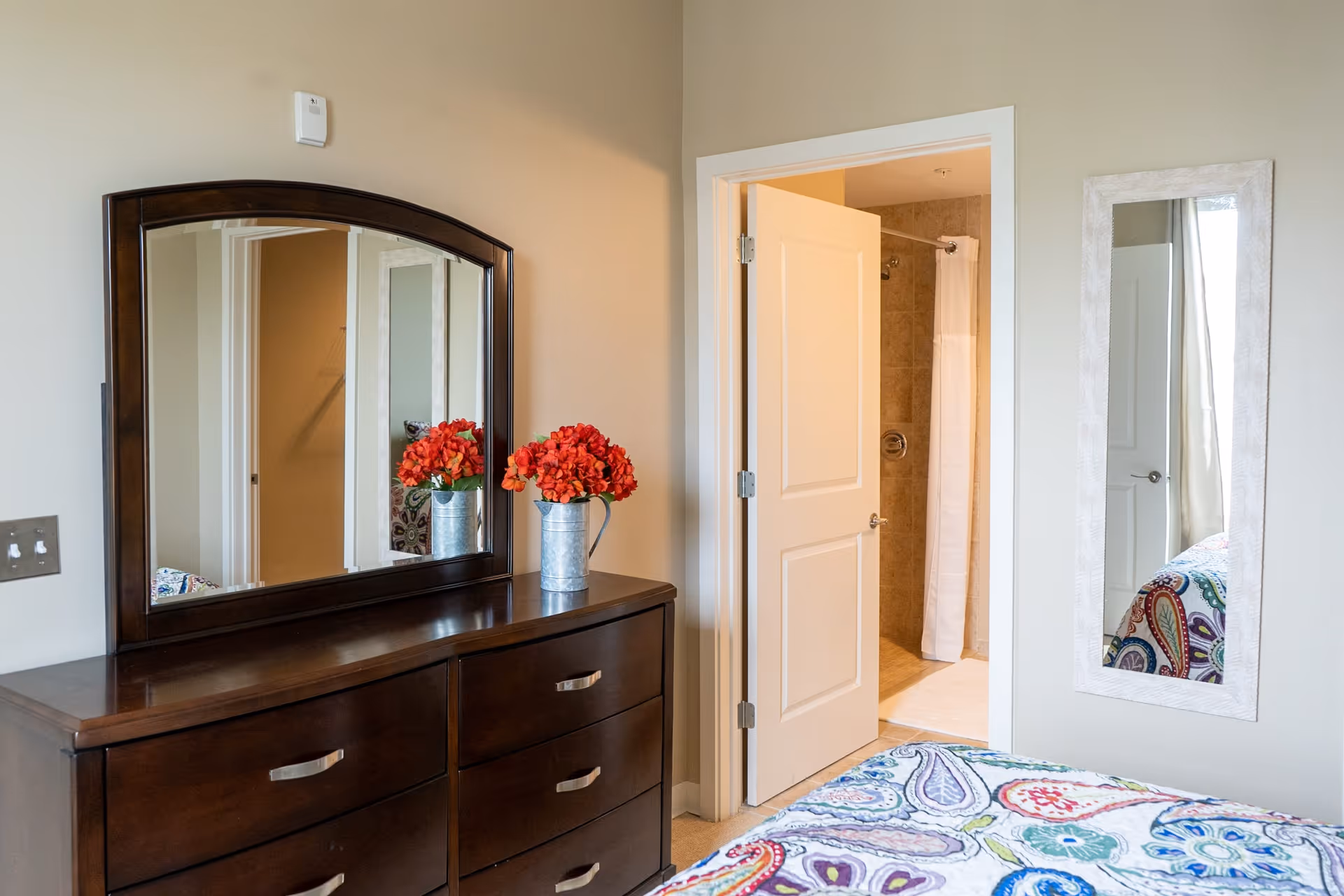 A bedroom interior featuring a dark wooden dresser with a large mirror and a vase of red flowers on top. To the right, an open door reveals a bathroom with a shower curtain. A colorful patterned bedspread is partially visible in the foreground, and a tall rectangular mirror is mounted on the wall beside the bathroom door.
