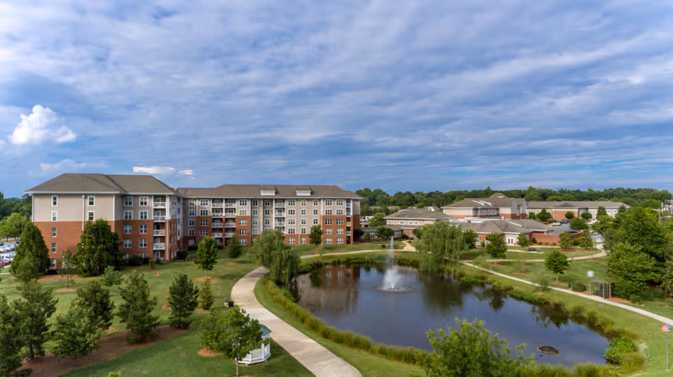 A large senior living facility complex with multiple connected buildings surrounding a landscaped area featuring a pond with a fountain, walking paths, and trees under a partly cloudy sky.