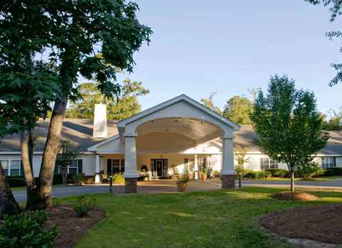 Front exterior view of a single-story senior living facility building with a covered entrance supported by columns, surrounded by trees and landscaped greenery under a clear sky.