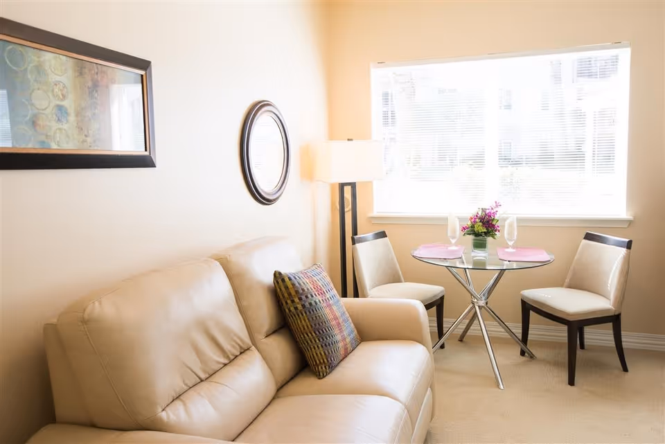 A cozy living area with a beige leather sofa adorned with a colorful pillow, a round glass table set with two chairs, two placemats, two glasses, and a small flower arrangement. A floor lamp stands next to the sofa, and a large window with blinds lets in natural light. The walls are decorated with a framed abstract painting and a round mirror.