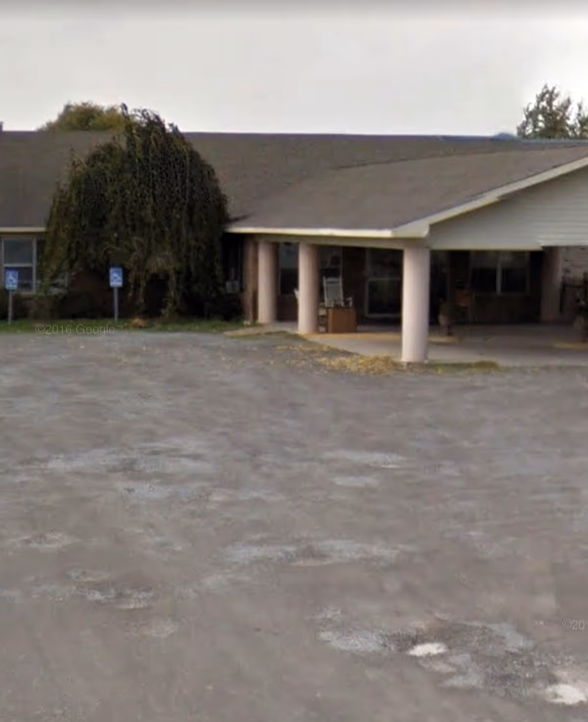 Front entrance of a single-story senior living building with a covered portico supported by columns and an adjacent parking area.
