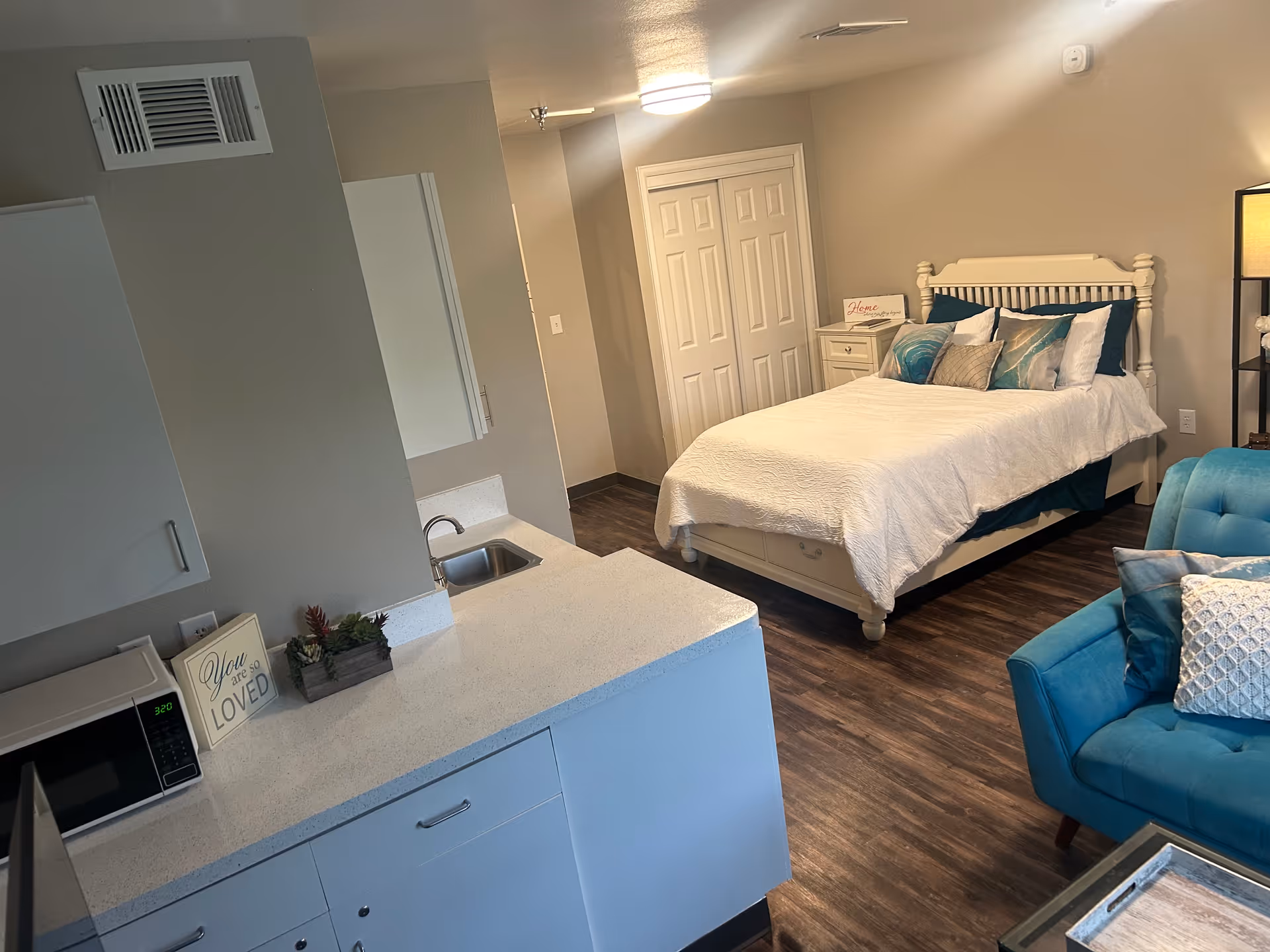 Interior view of a senior living facility room at Sodalis Texas City featuring a white bed with multiple pillows, a nightstand with a decorative sign, a blue armchair with cushions, and a kitchenette area with a microwave, sink, and countertop decorated with a small plant and a sign that reads 'You are so LOVED'. The room has wood flooring and neutral-colored walls.