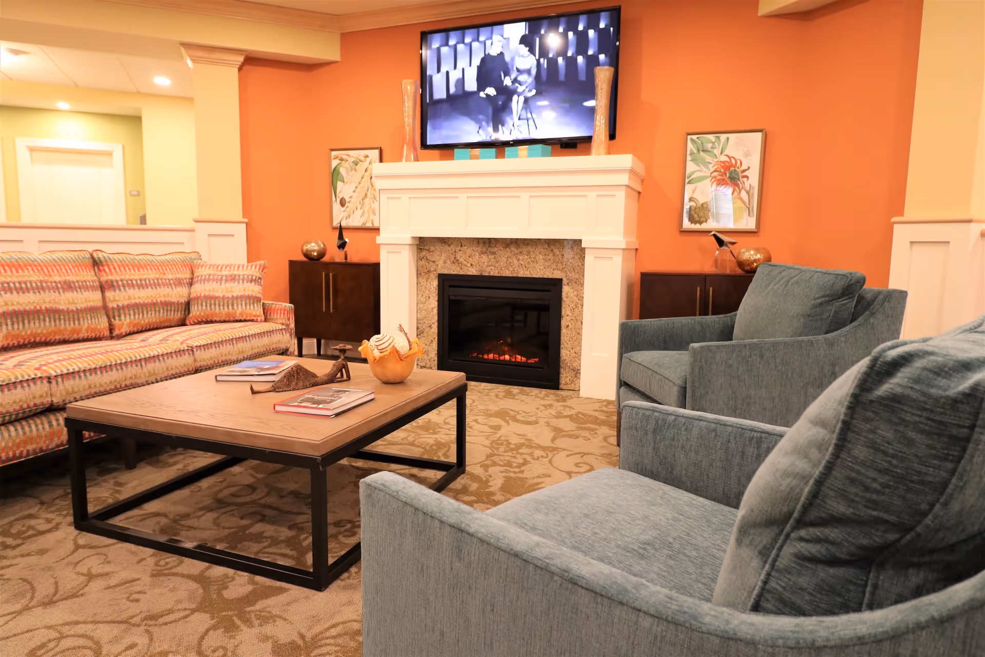 A cozy living room area with a patterned sofa, two blue armchairs, a wooden coffee table with books and decorative items, a fireplace with a white mantel, and a wall-mounted TV above the fireplace. The walls are painted orange and cream, and there are framed artworks on either side of the fireplace.