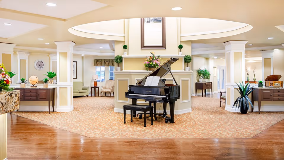 A spacious and elegant common area in a senior living facility featuring a black grand piano with an open music book on its stand, surrounded by beige walls with white trim and decorative columns. The room has patterned carpet flooring with a wooden floor section in the foreground. There are various plants, floral arrangements, and comfortable seating areas with armchairs and side tables visible in the background. The ceiling has recessed lighting and a large circular skylight above the piano.