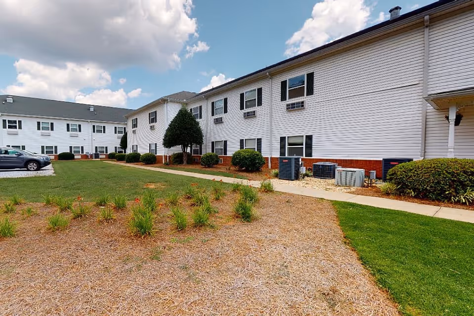 Two-story white senior living building with a courtyard, walkway, and landscaped lawn under a partly cloudy sky.