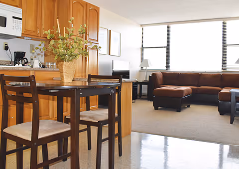 Interior view of a senior living facility apartment at Irving Cypen Tower featuring a small dining area with a dark wooden table and two chairs, a kitchen with wooden cabinets, a microwave, and a coffee maker, and a living room with a brown sectional sofa, a TV on a stand, and large windows with blinds letting in natural light.