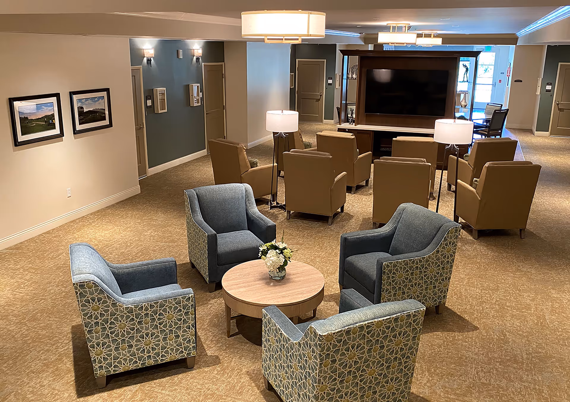 A cozy senior living facility common area with four patterned blue armchairs arranged around a round wooden table with a small flower arrangement. Behind them are two rows of tan armchairs facing a large flat-screen TV mounted on a wooden entertainment unit. The room has beige carpet, soft lighting from ceiling fixtures and lamps, and framed landscape pictures on the wall.