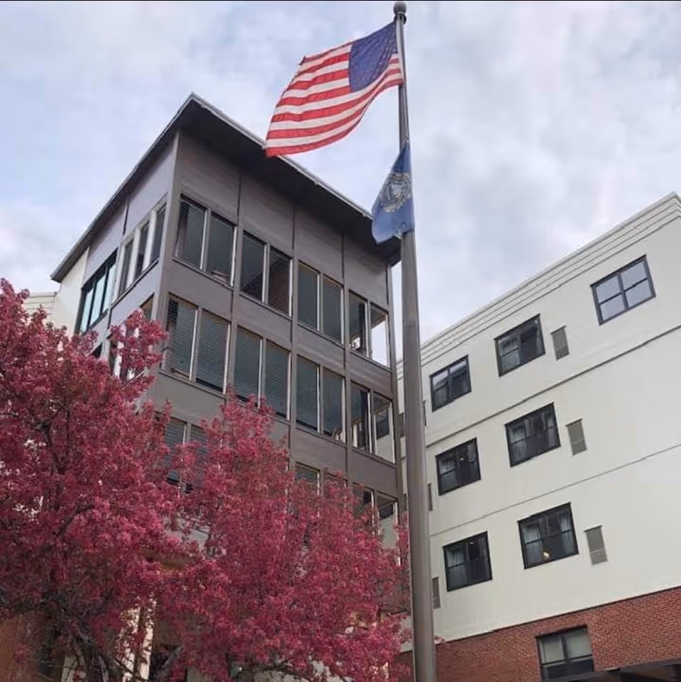 Exterior view of a multi-story building with large windows, an American flag and another flag on a flagpole in front, and pink flowering trees in the foreground under a cloudy sky.