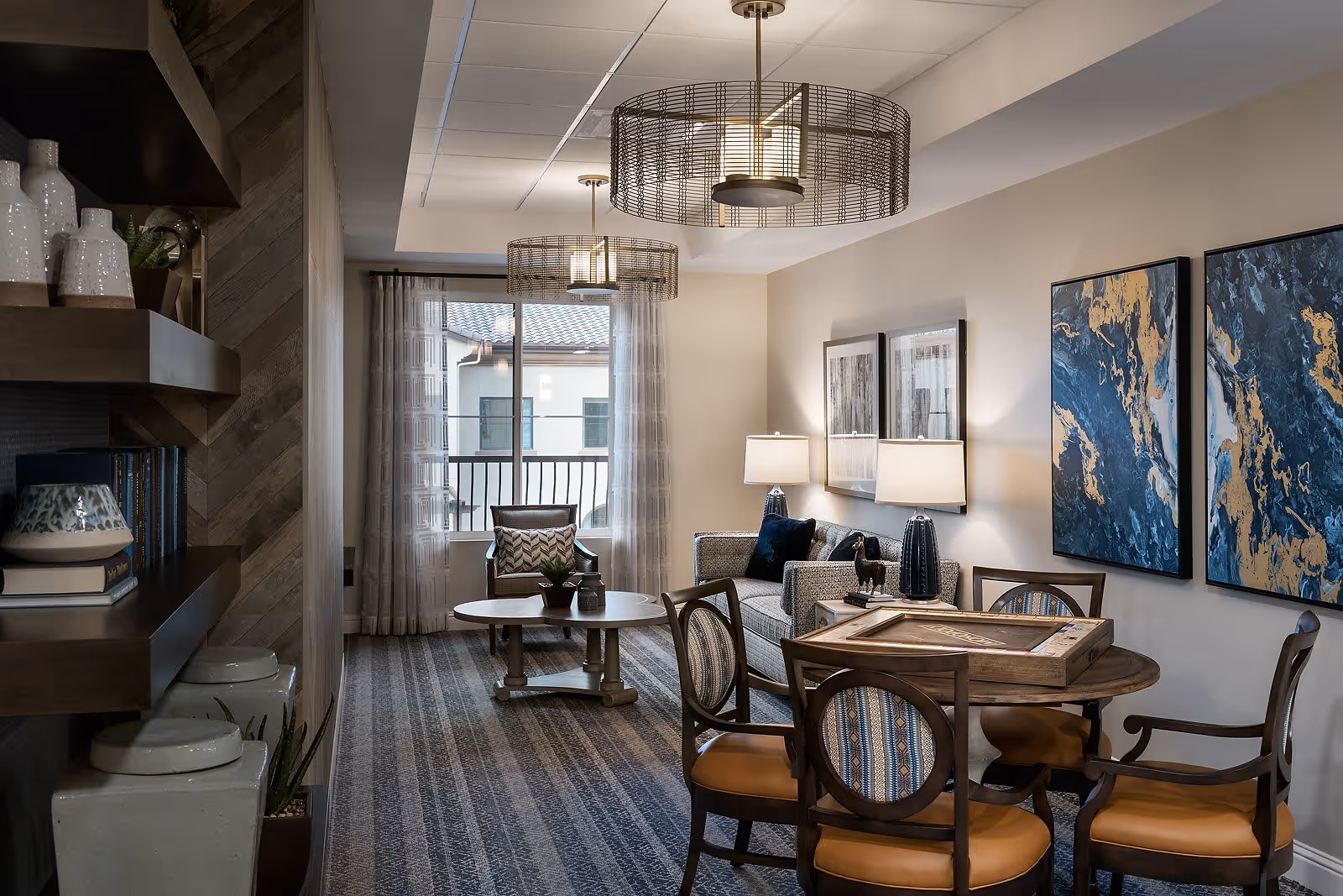 A cozy living room area with a round wooden table and four chairs with patterned upholstery. Behind the table is a gray sofa with two lamps on side tables and two framed artworks on the wall. A coffee table and an armchair are positioned near a window with sheer curtains, allowing natural light to enter. The room features modern pendant lights and a carpeted floor with a striped pattern.