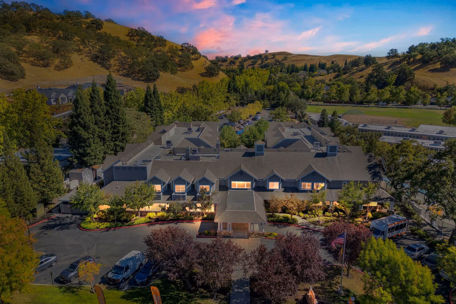 Aerial view of a senior living facility named Sunrise of Danville surrounded by trees and hills during sunset, with a parking area and an American flag in front of the building.