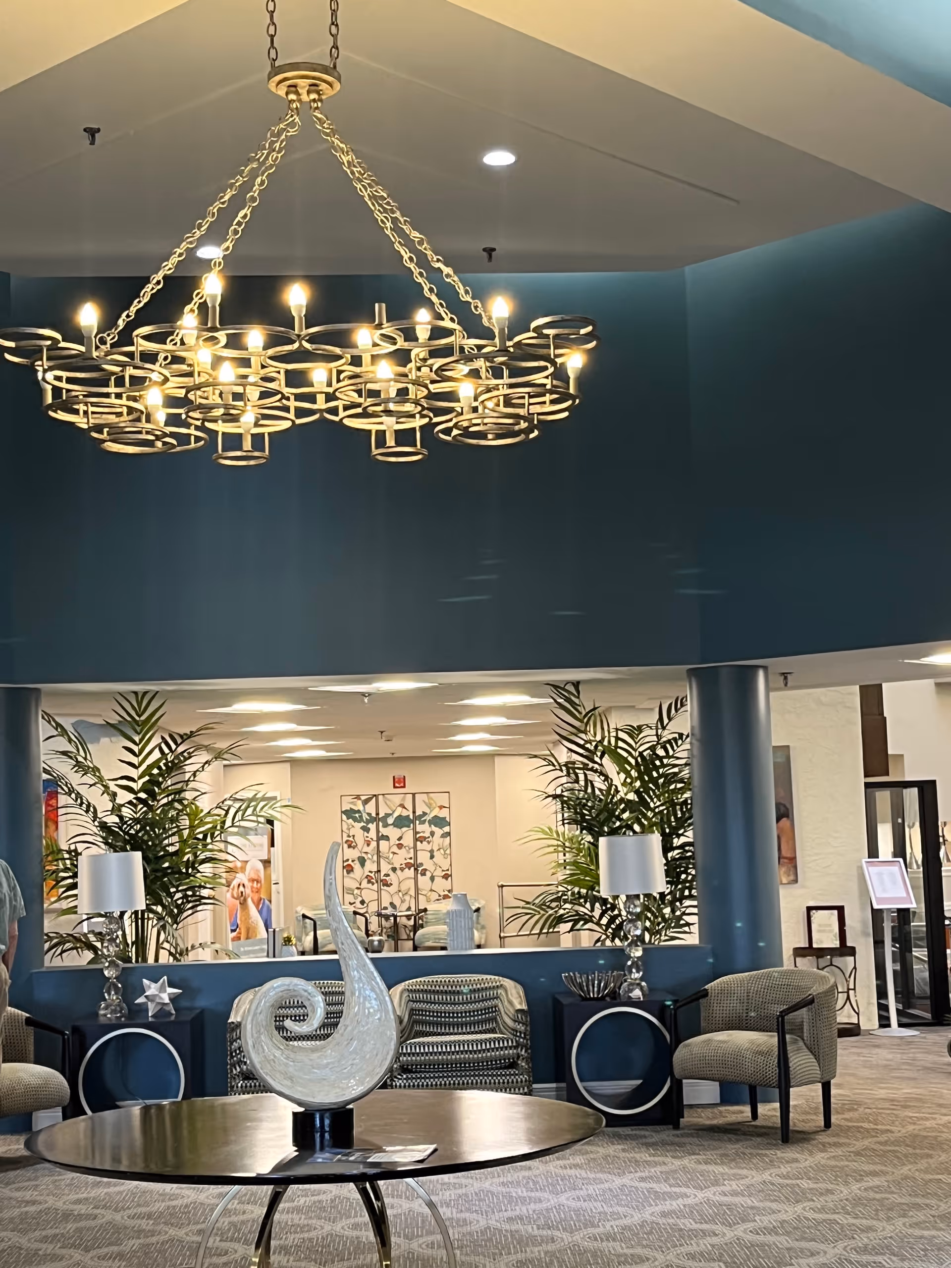 A spacious senior living community lobby with a modern chandelier hanging from the ceiling. The room features a round table with a decorative glass sculpture in the center, surrounded by comfortable chairs and side tables with lamps. Large plants and a mirror wall add to the welcoming atmosphere.