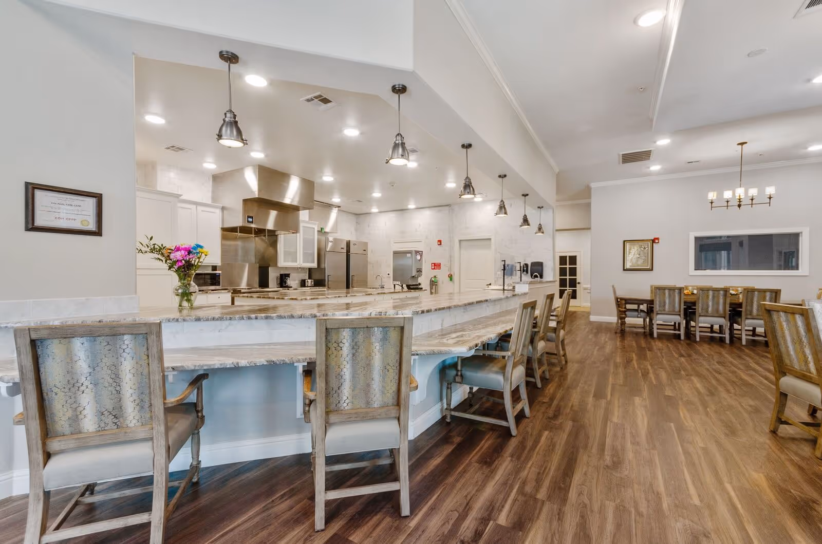 A spacious dining area with a long marble countertop bar lined with wooden chairs with patterned backs. The kitchen area behind the bar features stainless steel appliances, white cabinetry, and pendant lighting. In the background, there are additional dining tables and chairs on a wood floor, with neutral-colored walls and ceiling lights.