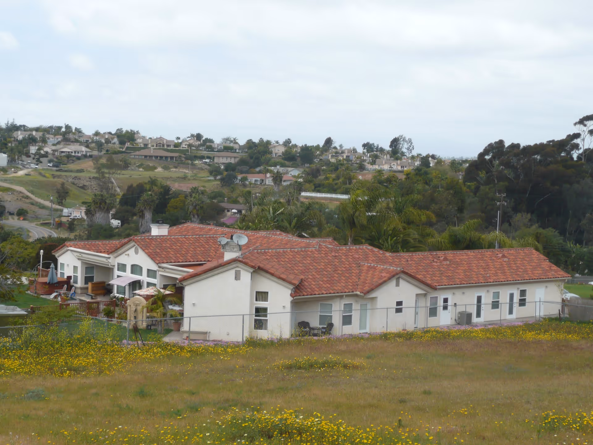View of a single-story building with a red tiled roof surrounded by a grassy field with yellow flowers. The building is located in a suburban area with other houses and trees in the background under a cloudy sky.