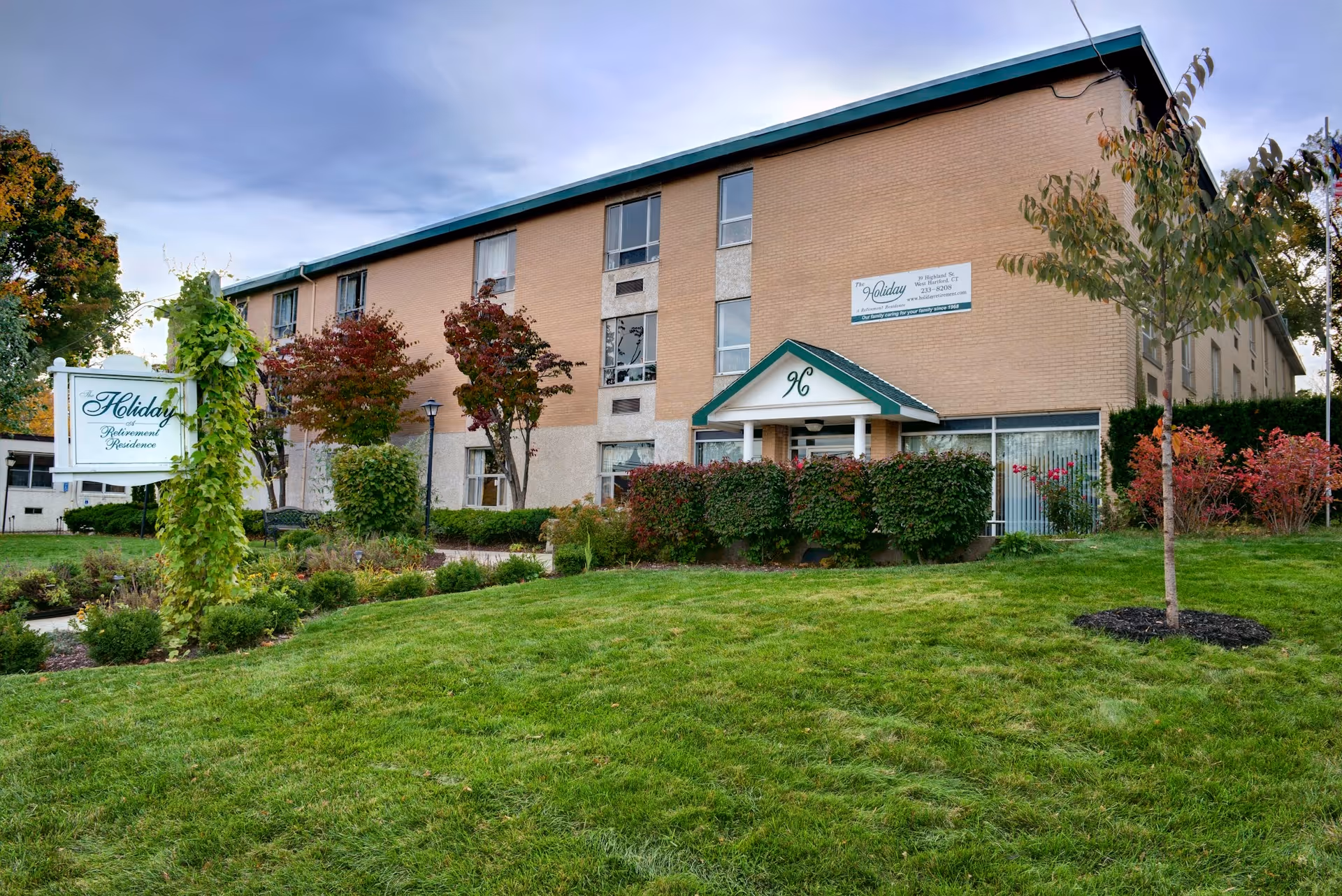 Exterior view of a three-story senior living facility building with a green lawn and landscaped bushes and trees in front. A white sign near the entrance reads 'Holiday Retirement Residence.' The building has a beige and light brown facade with several windows and a small covered entrance with the number 96 above it.