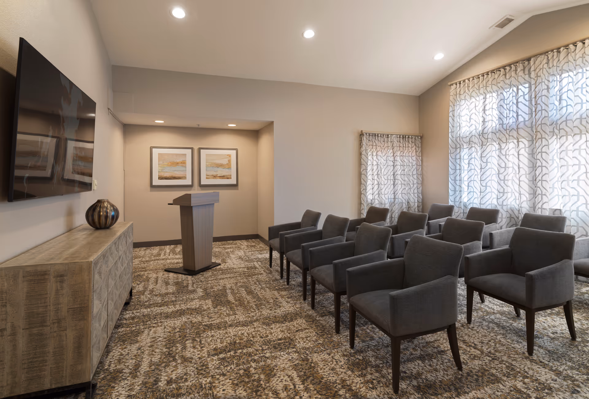 A small conference or meeting room with rows of gray upholstered chairs facing a wooden podium. The room has patterned carpet, light beige walls, two framed abstract paintings on the far wall, a large flat-screen TV mounted on the left wall, and windows with sheer patterned curtains on the right side.