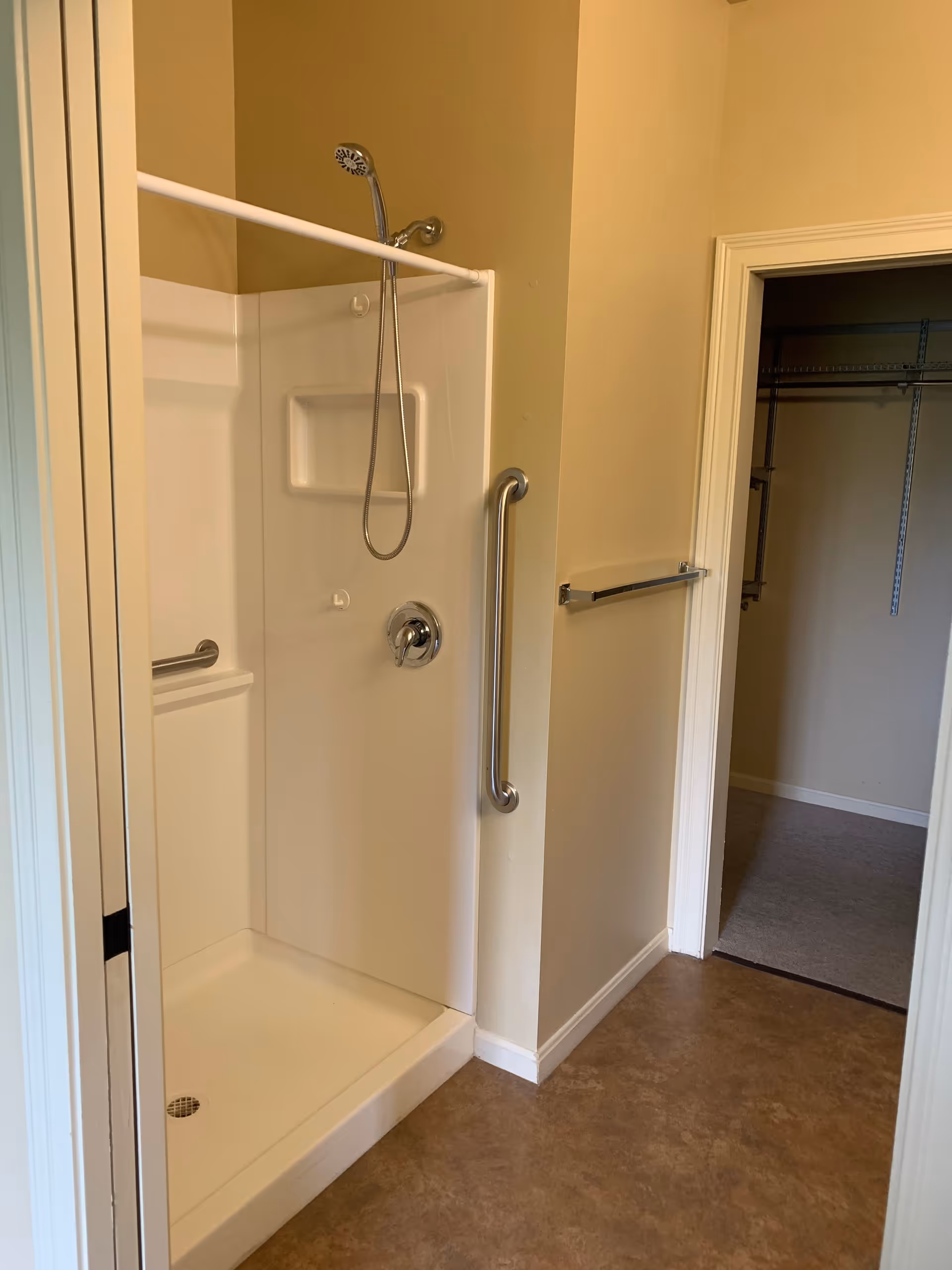 A clean, beige-toned bathroom featuring a white walk-in shower with a handheld showerhead, grab bars for safety, and a built-in soap dish. Adjacent to the shower is a towel rack mounted on the wall, and an open doorway leads to a carpeted closet area with shelving.