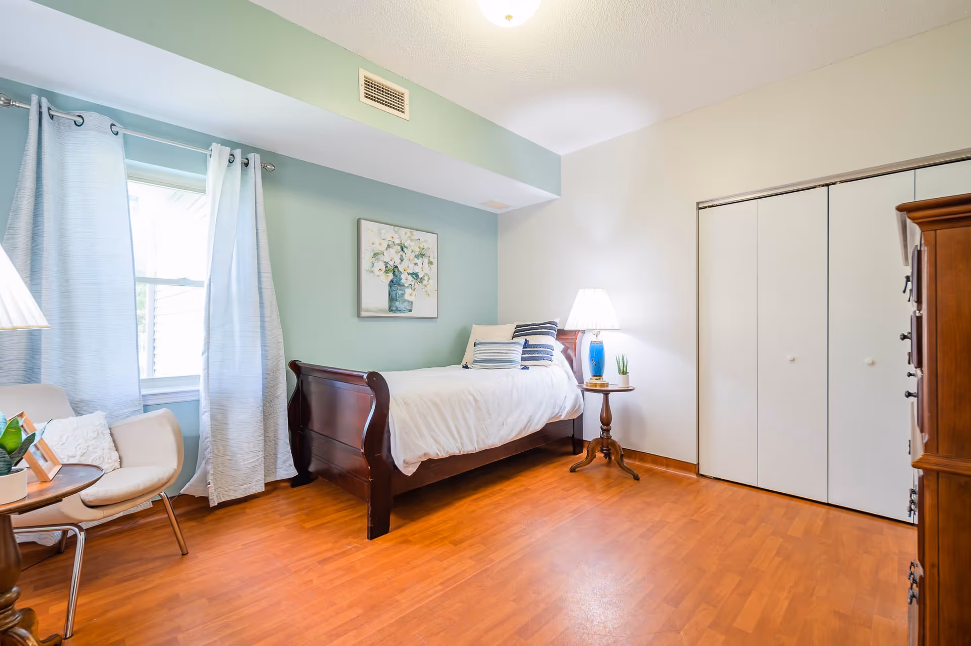 A bright bedroom with a wooden single bed against a light green wall, decorated with white and blue pillows. There is a small round wooden side table with a blue lamp and a small plant next to the bed. A window with light blue curtains is on the left side, and a beige chair with a cushion is nearby. The floor is wooden, and there is a white closet with sliding doors on the right side.